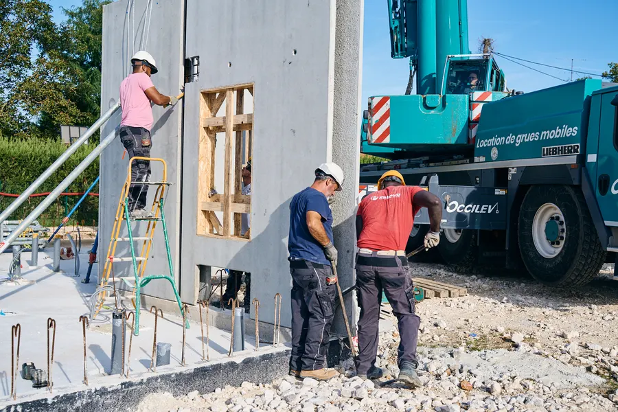 Compagnons au pied d'un mur préfabriqué en cours de pose, grue mobile derrière